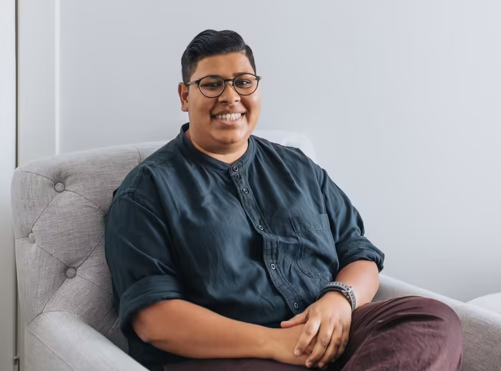 Jaini Shah sitting on a couch smiling with a navy blue shirt on and maroon pants. Jaini is wearing round glasses and smiling at the camera.